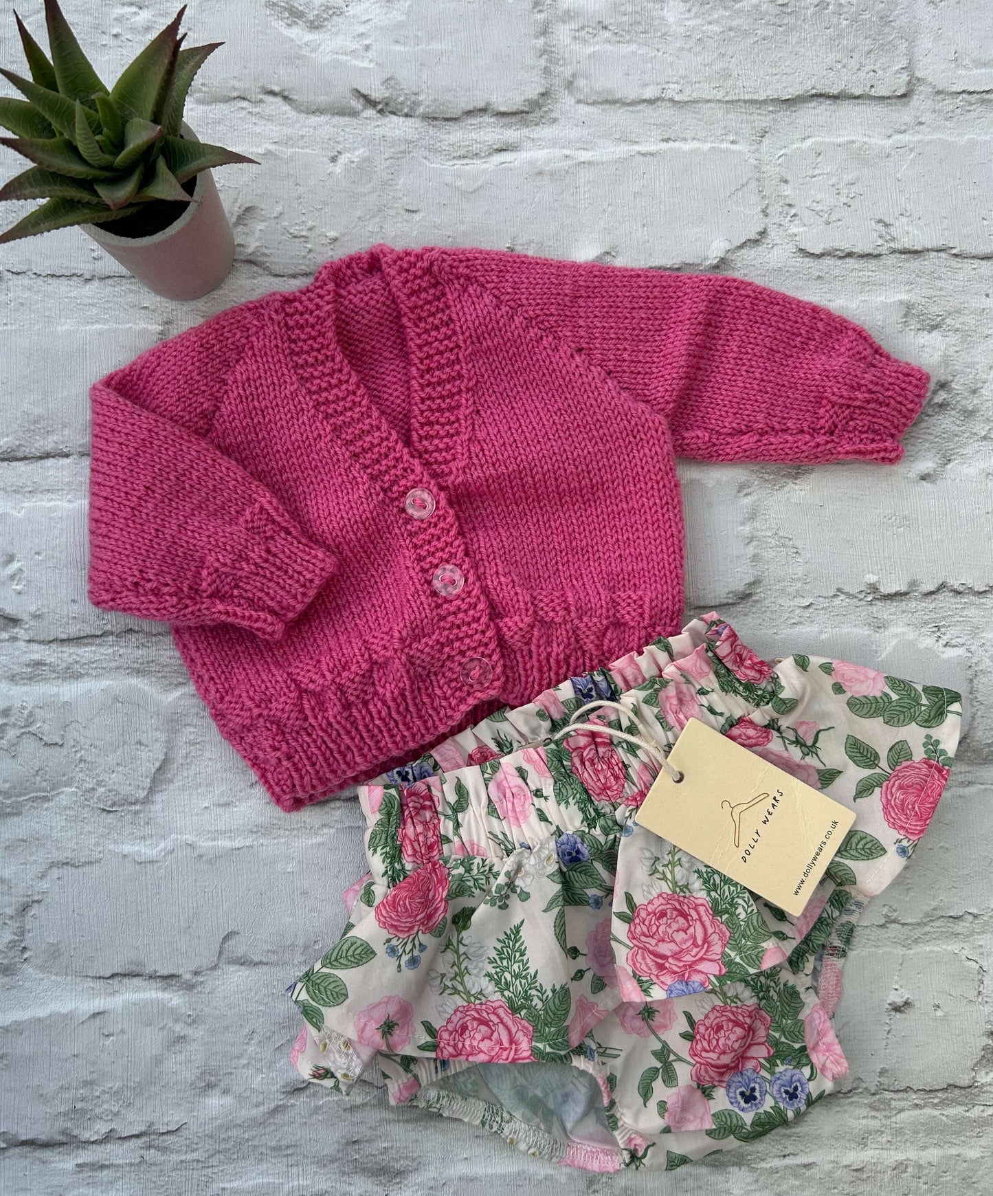 Pink knitted baby hand knitted cardigan to personalise with outfit of floral-patterned baby bloomers on a light gray surface.