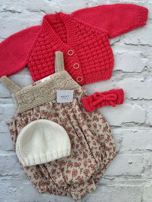 Baby outfit set with ready to personalise hand knitted coral baby cardigan floral cotton romper, white hat, and coral headband on a light gray background.