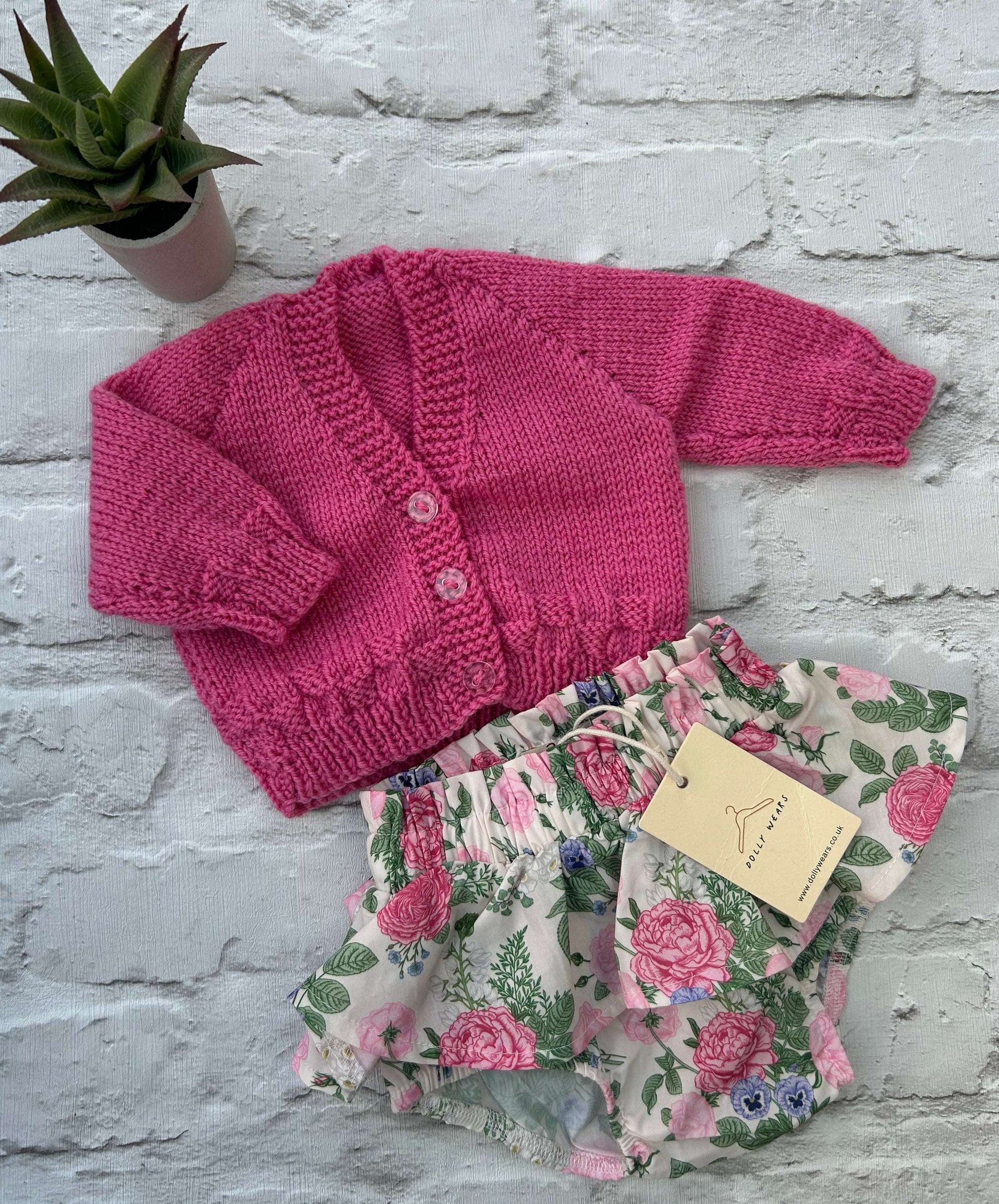 Pink knitted baby hand knitted cardigan to personalise with outfit of floral-patterned baby bloomers on a light gray surface.