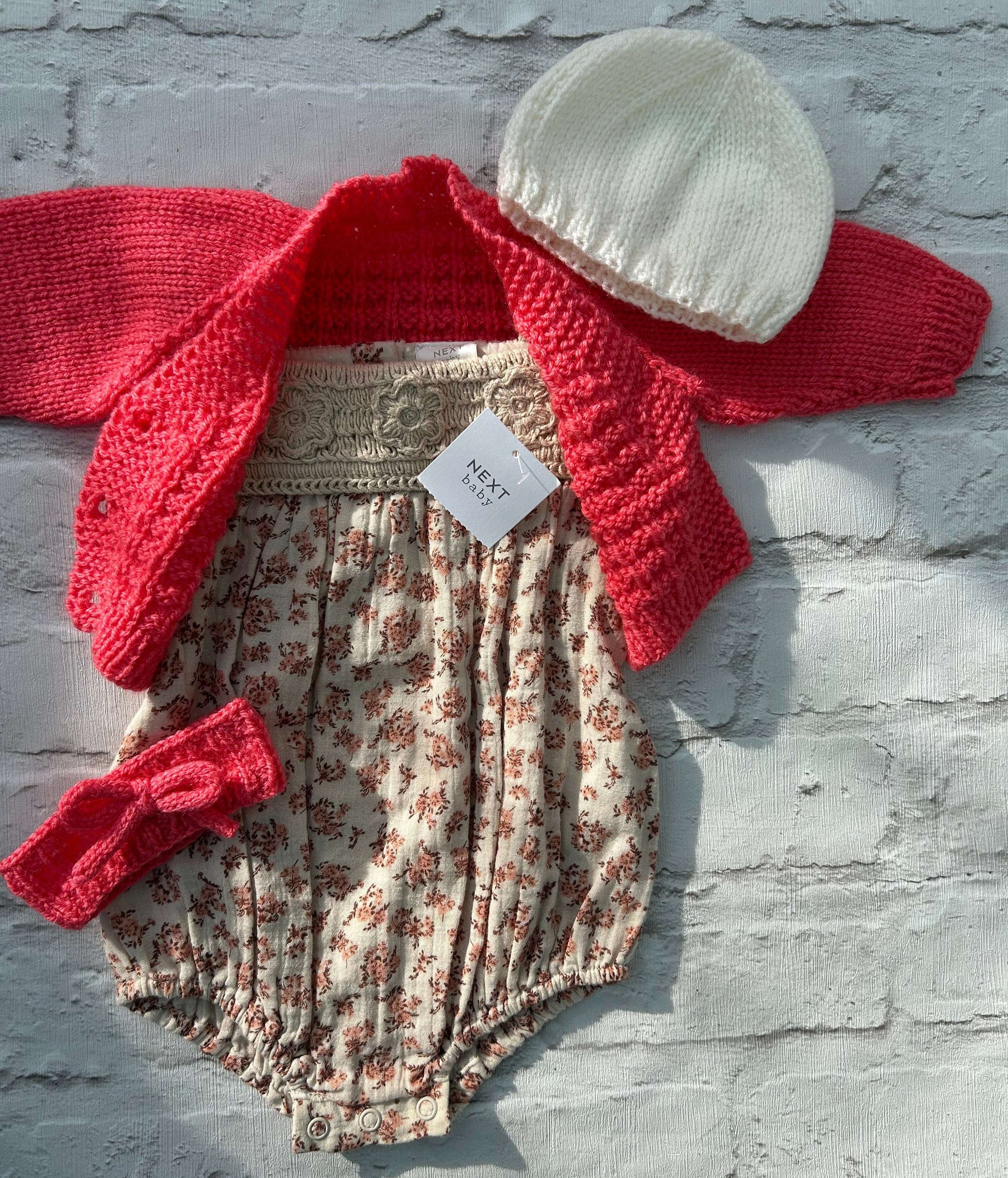 Baby outfit set with ready to personalise hand knitted coral baby cardigan floral cotton romper, white hat, and coral headband on a light gray background. size 0-3 months 