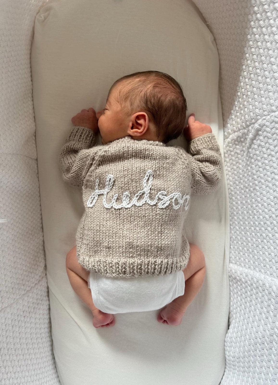 Newborn baby in a crib wearing a hand knitted baby cardigan in beige with hand embroidered with baby name 'Hudson' on it in white embroidery yarn