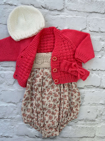 Baby outfit set with ready to personalise hand knitted coral baby cardigan floral cotton romper, white hat, and coral headband on a light gray background.