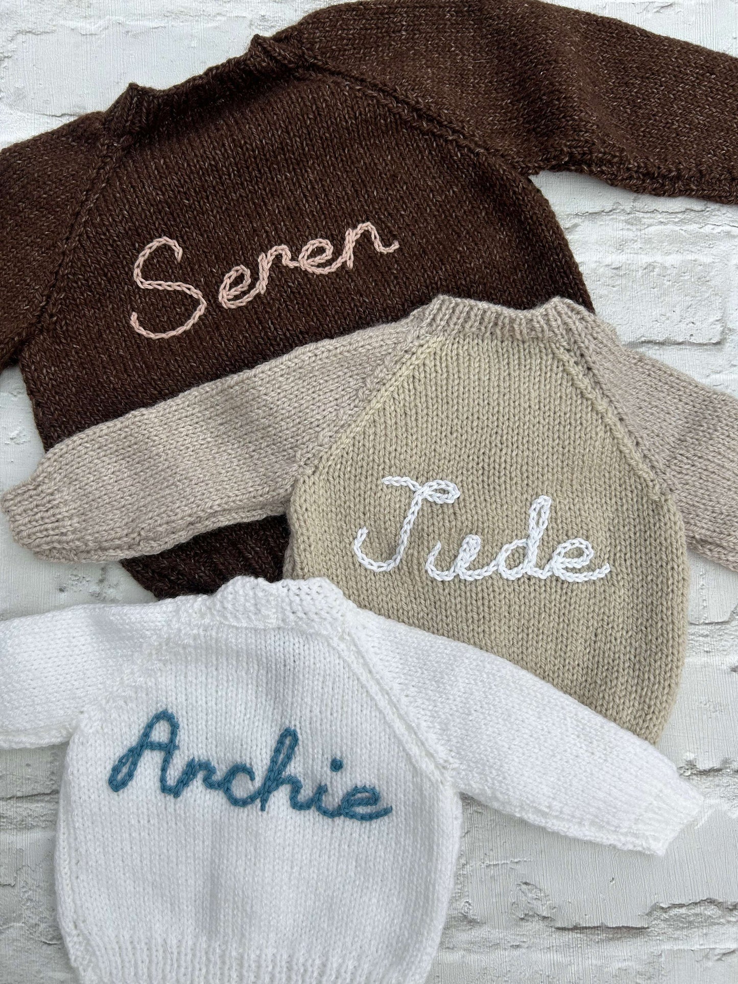 Selection of personalised hand knitted baby cardigans laid flat showing embroidered names in various colours | Little Name Knits

