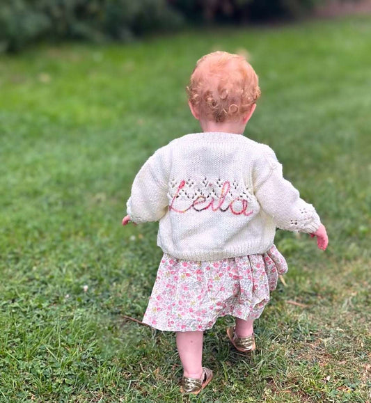 Child wearing a white personalised hand knitted cardigan with 'Leila' embroidered on it, standing on grass.