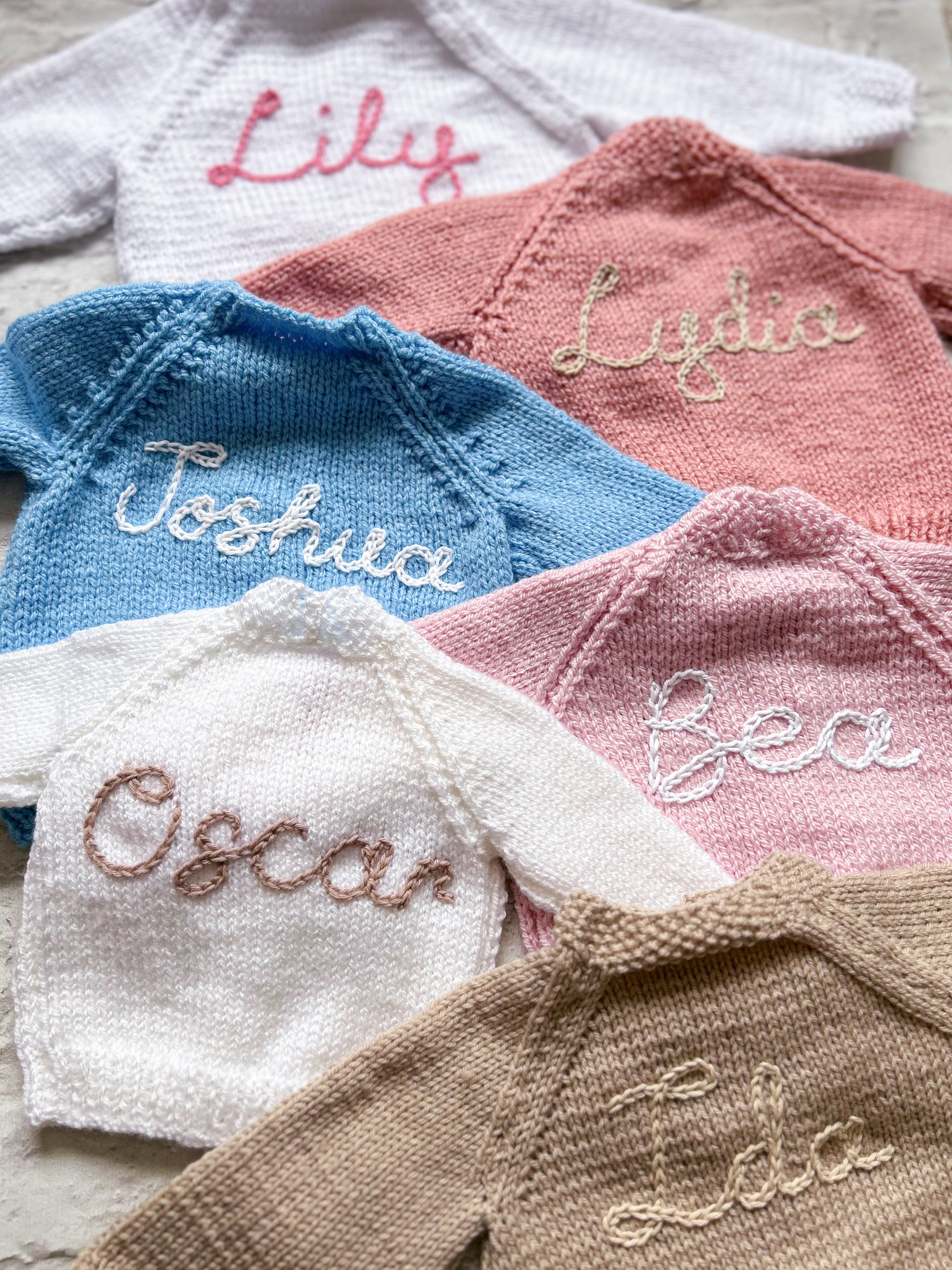 Collection of hand made hand knitted baby sweaters with hand embroidered baby names on a light background