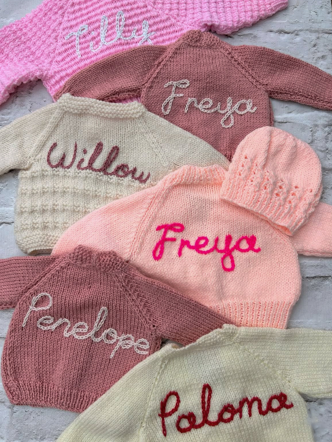 Collection of hand knitted cardigans and a hat with hand embroidered names. Pretty pink colours, various styles