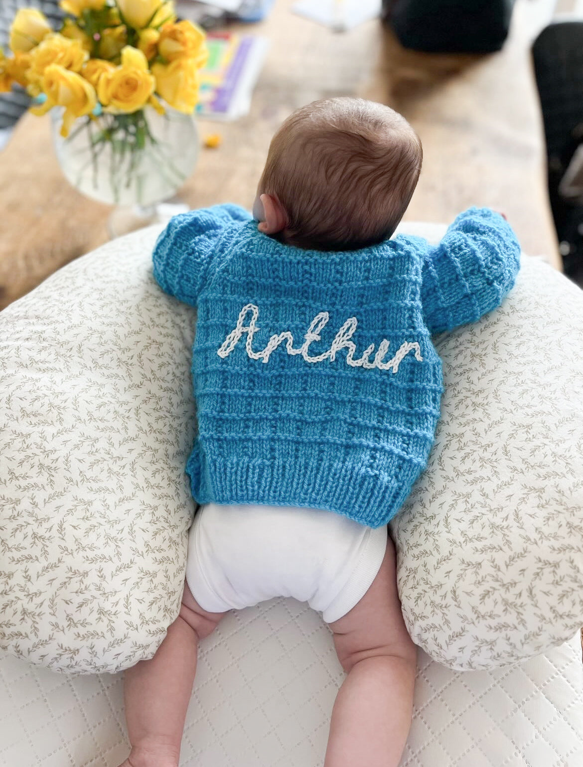 Baby wearing a blue knitted cardigan with 'Arthur' on it, sitting on a textured surface. Example of previously sold hand knitted cardigan featured on a Baby 