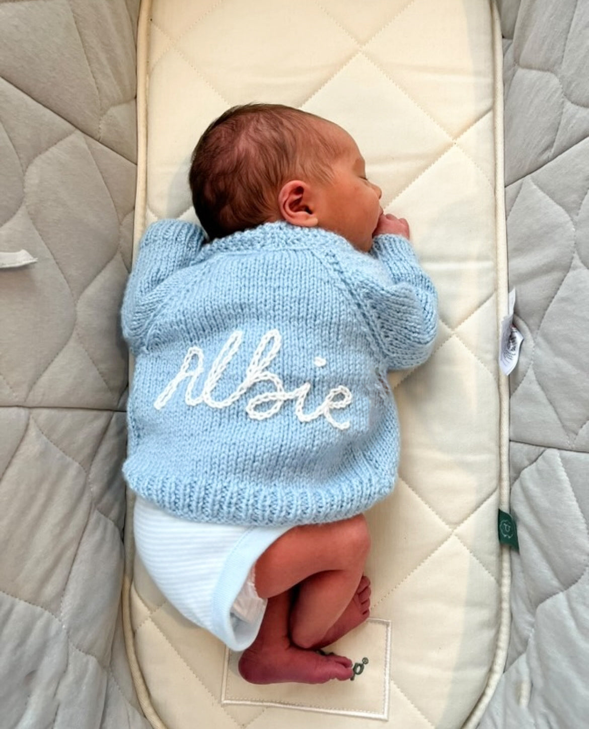 Newborn baby wearing a baby blue hand knitted cardigan hand embroidered with 'Albie' lying on a quilted surface in moses basket