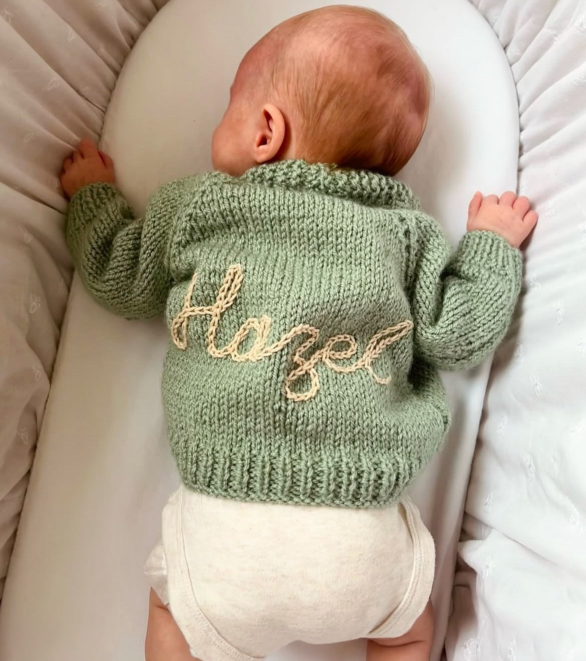 Example of previous work Baby wearing a green hand knitted sweater with 'Hazel' hand embroidered on it, laying in a white crib.