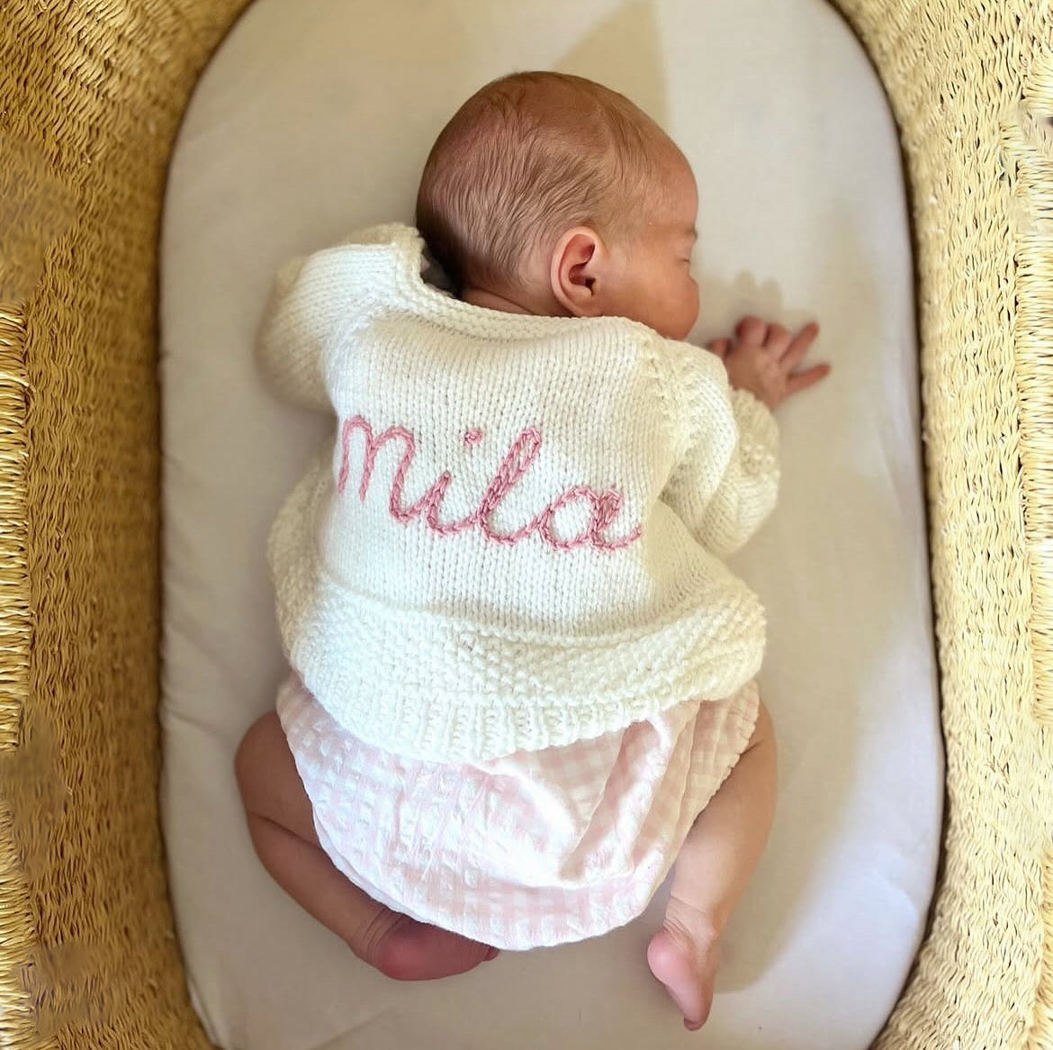 Baby in a cream hand knitted knitted outfit with 'Mila' hand embroidered on it in rose pink, lying in a moses basket.